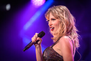 A performer, Taylor Swift, singing into a microphone on stage with colorful stage lighting in the background in Europe.